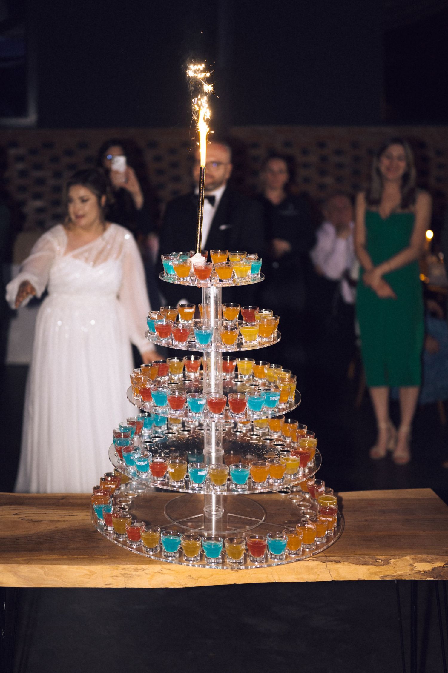 Multi-tiered shot glass tower display with colorful drinks and a sparkler at a wedding reception.