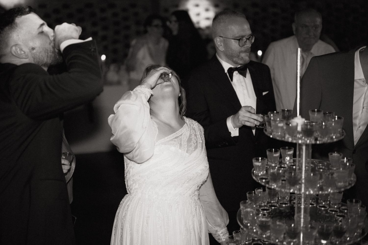 Black and white photo of bride and groom doing shots at wedding reception near champagne tower.