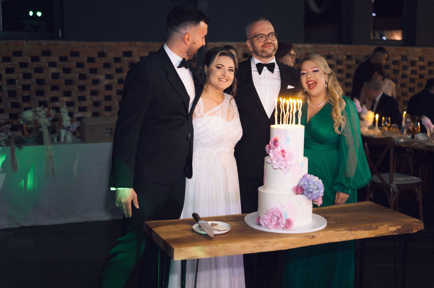 Wedding couple poses with guests beside elegant white tiered cake with candles and floral decorations.