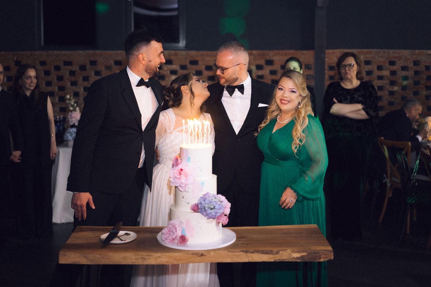 Wedding couple cutting a white floral cake with guests in formal attire at an elegant indoor reception.