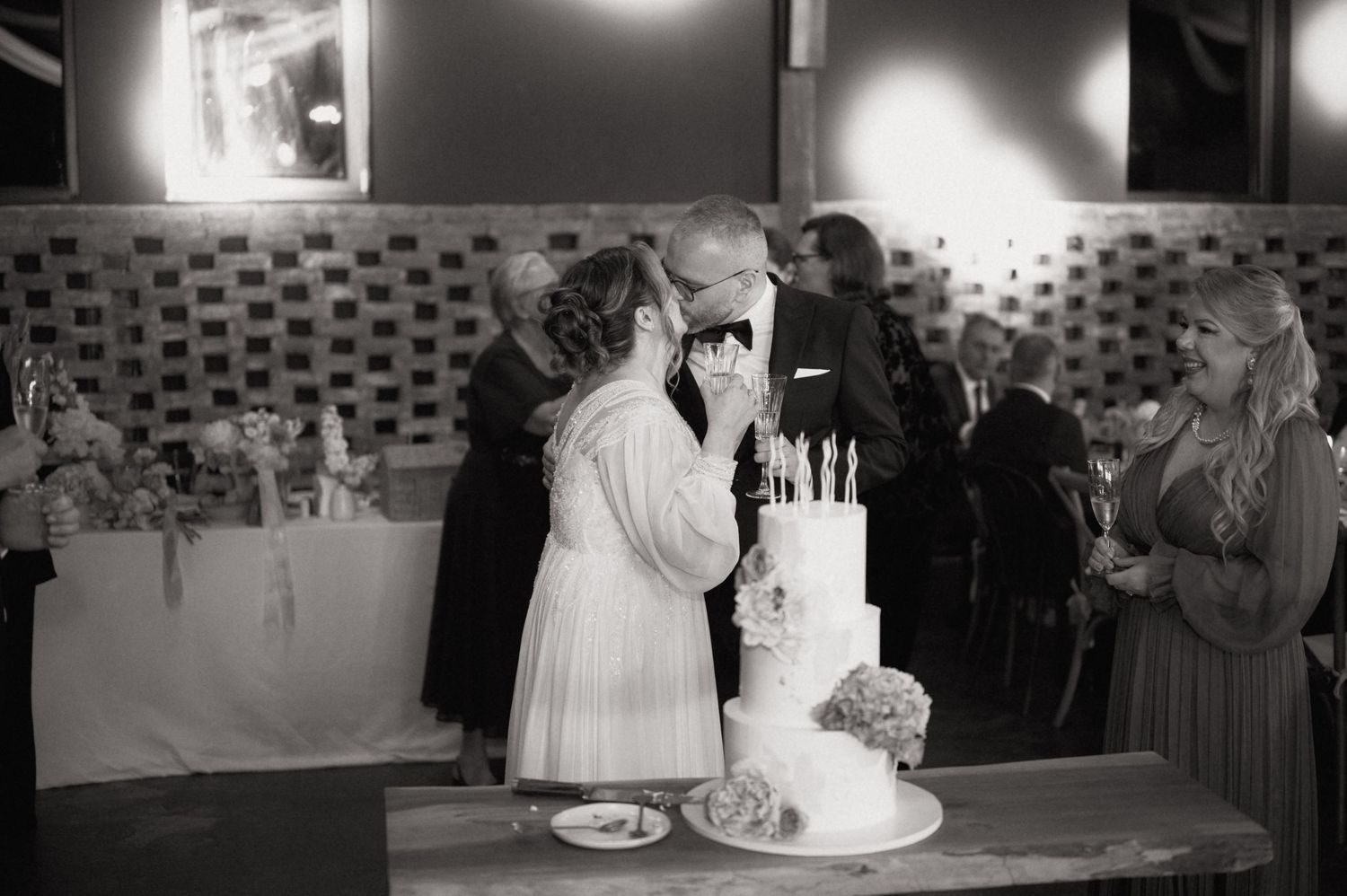 Black and white photo of a couple sharing a kiss while cutting their elegant wedding cake at their reception.