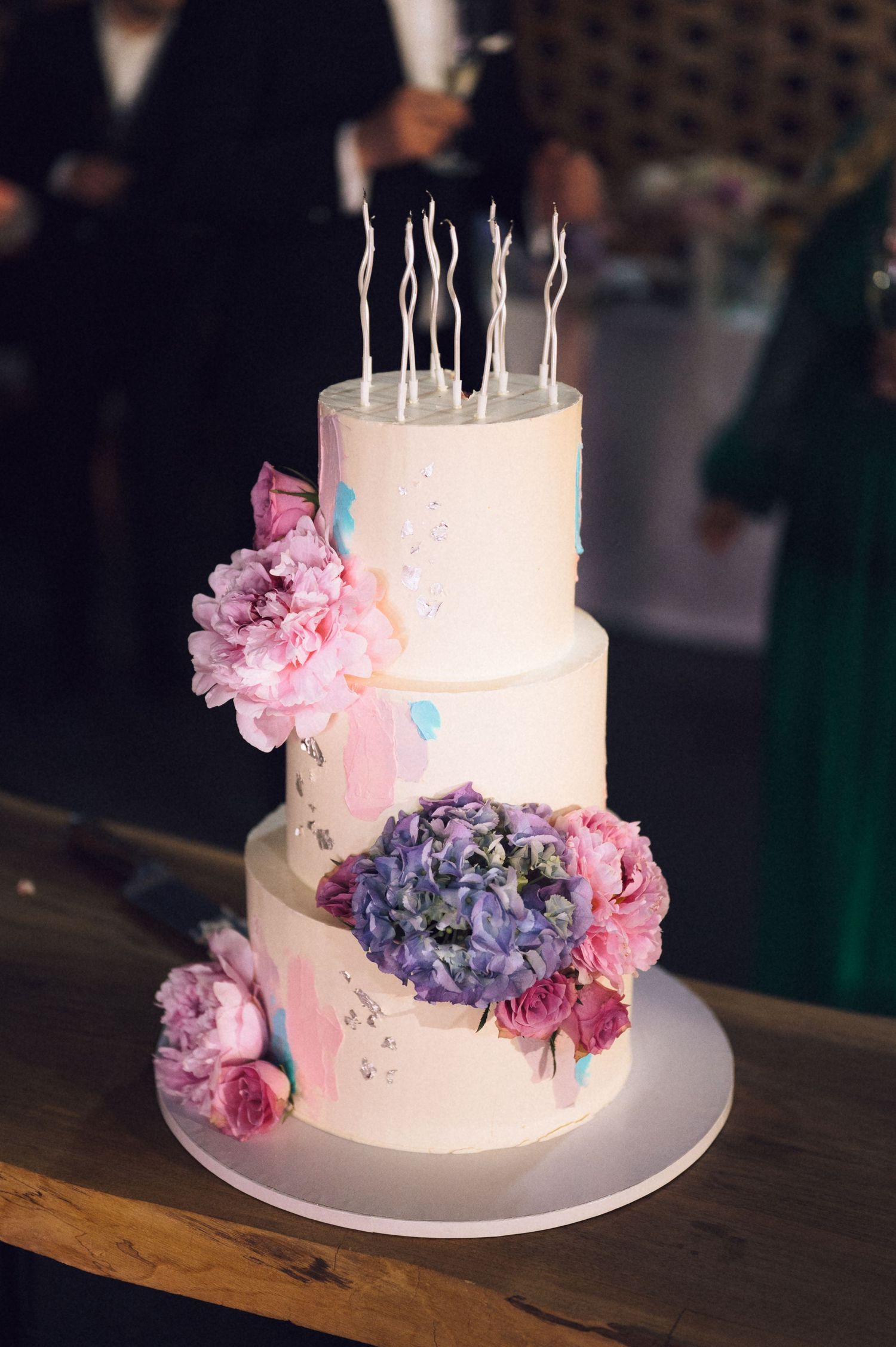 Three-tier white wedding cake with pink peonies, purple hydrangeas, watercolor brushstrokes, and decorative candles on wooden table.