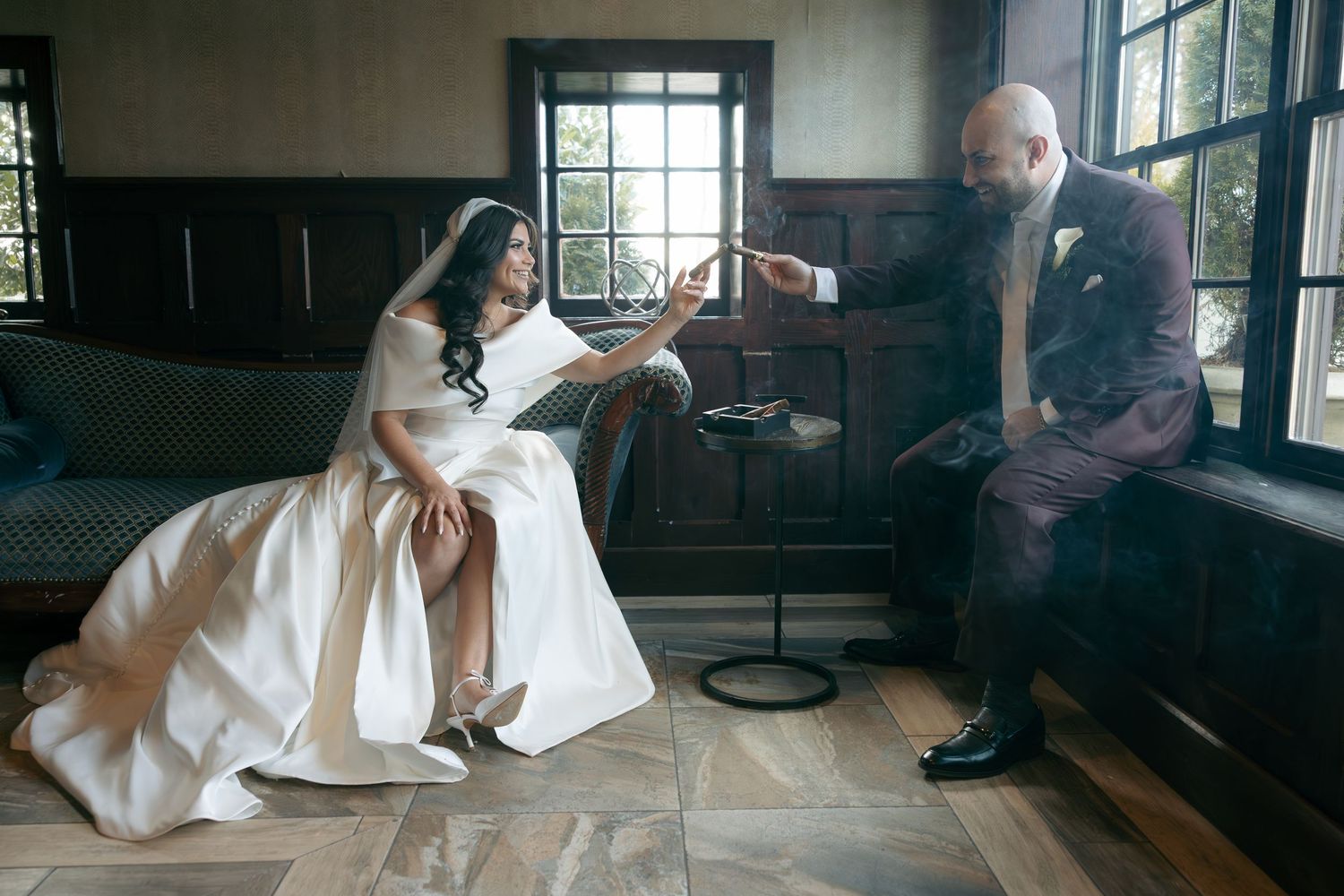 Bride in white gown and groom in burgundy suit share an intimate moment seated together in an elegant wood-paneled room.