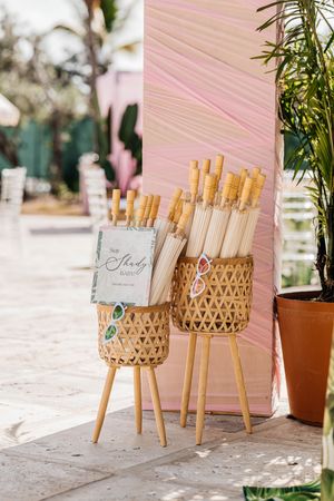 Wicker baskets holding white parasols with bamboo handles displayed at an outdoor wedding ceremony entrance with pink backdrop.