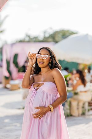 Pregnant woman in pink dress and cat-eye sunglasses smiling outdoors at a sunny event.