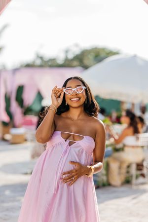 Smiling pregnant woman in pink strapless dress holding cat-eye sunglasses at an outdoor event.