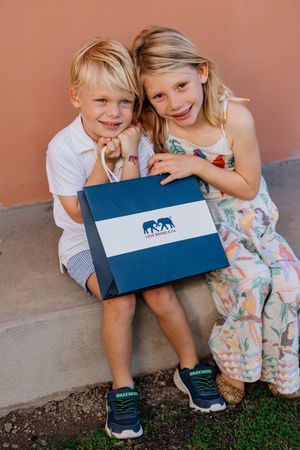 Two smiling children sitting together holding a navy blue gift bag with an elephant logo.