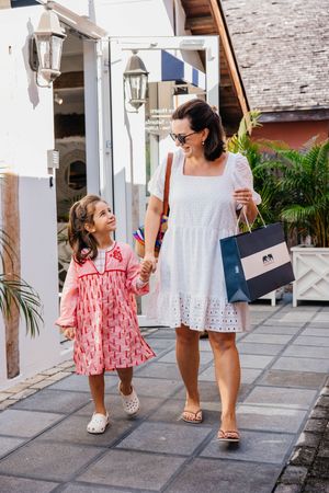 A woman in a white dress and a young girl in pink walk hand-in-hand while shopping outdoors.