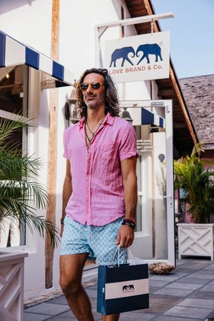 Man in pink shirt and blue shorts carrying a Love Brand & Co. shopping bag outside the store.