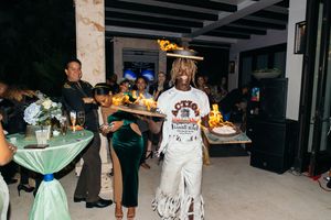 A performer in a wide-brim hat and fringed outfit carries a tray at an elegant evening party.