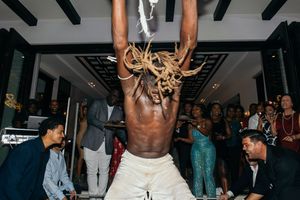 A shirtless performer with dreadlocks entertains a crowd at an indoor event, hanging upside down dramatically.