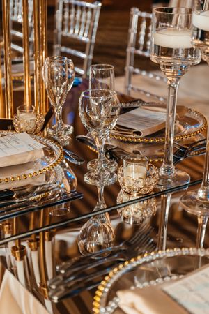 Elegant wedding reception table with crystal glassware, gold charger plates, and candles on mirrored surface.