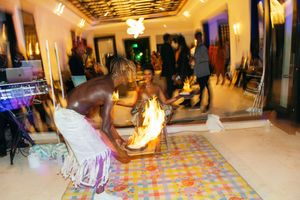 Fire dancer performing at an indoor cultural event, wearing a white sarong, with flames on a colorful mat.
