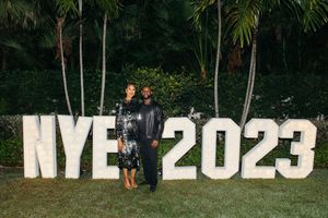 Couple poses in front of illuminated NYE 2023 sign surrounded by palm trees at New Years Eve celebration.