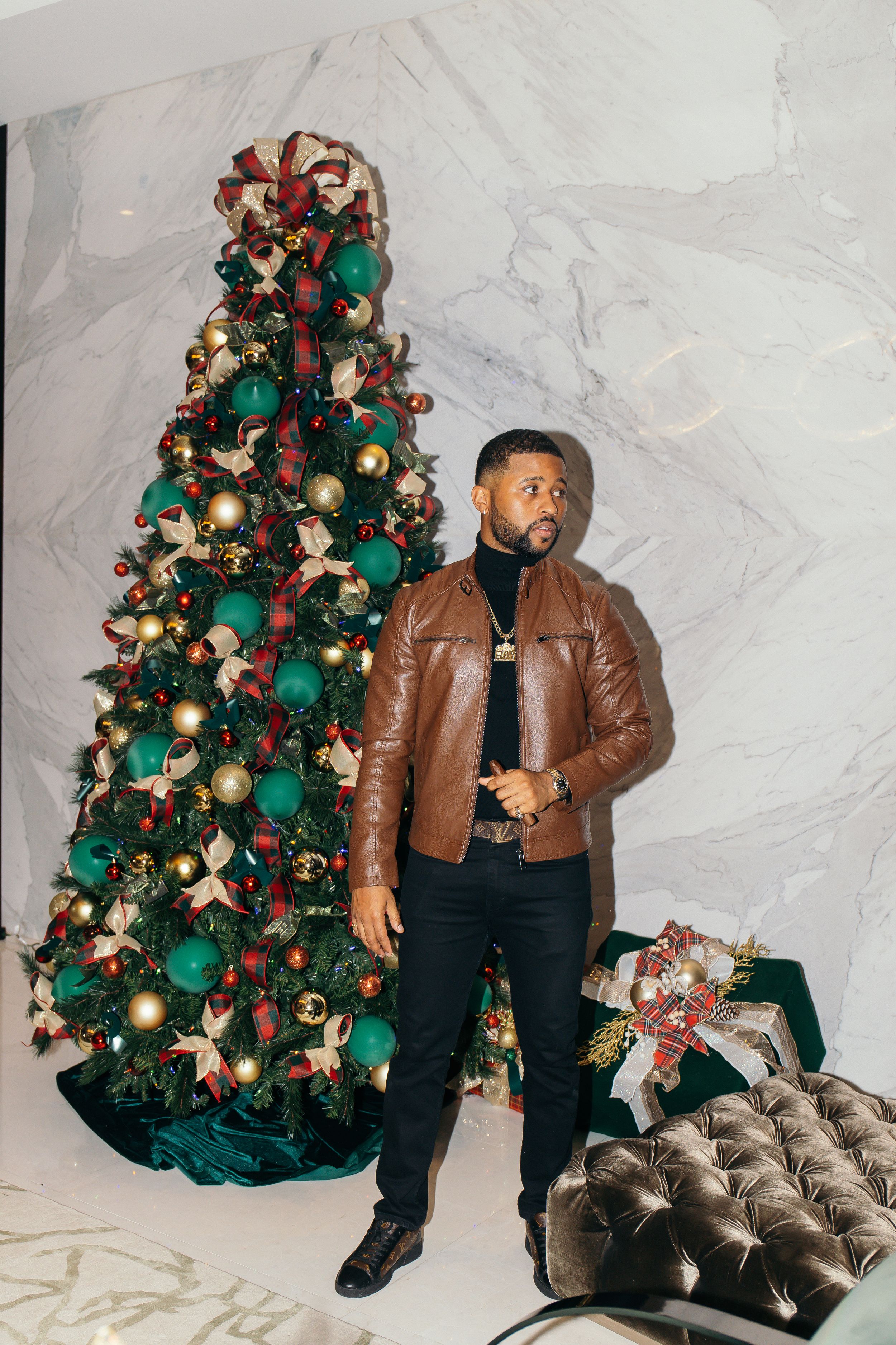 Man in brown leather jacket standing beside tall decorated Christmas tree with green and gold ornaments.