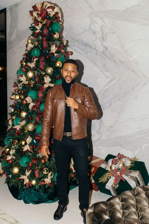 Man in brown leather jacket poses beside decorated Christmas tree with marble wall background.