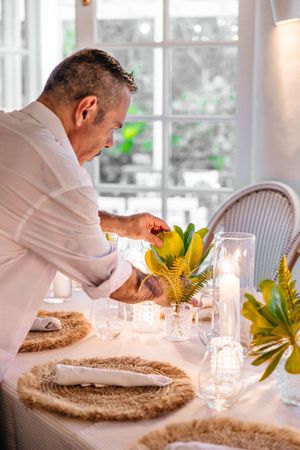 Man in white shirt arranging floral centerpiece on elegantly set dining table with candles and woven placemats.