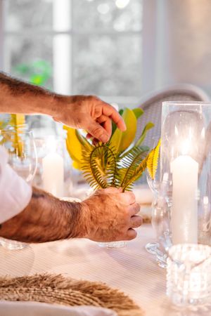 Man arranging tropical yellow fern leaves in glass vase on elegant table setting with white candles.