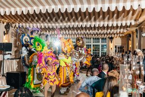 Colorful Mardi Gras performers in elaborate costumes dance at an elegant indoor event venue with striped awning.