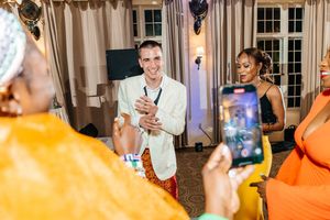 Man in white blazer clapping and smiling at a festive indoor gathering with guests in colorful attire.