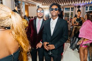 Two stylish men in suits and sunglasses pose at an elegant party event.