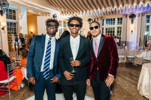 Three stylish young men in formal suits and sunglasses pose together at an elegant indoor event venue.