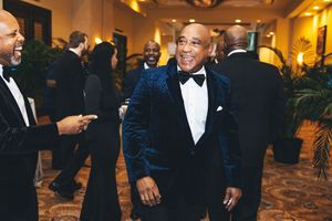 Man in blue velvet tuxedo jacket smiling at formal gala event surrounded by guests in black tie attire.