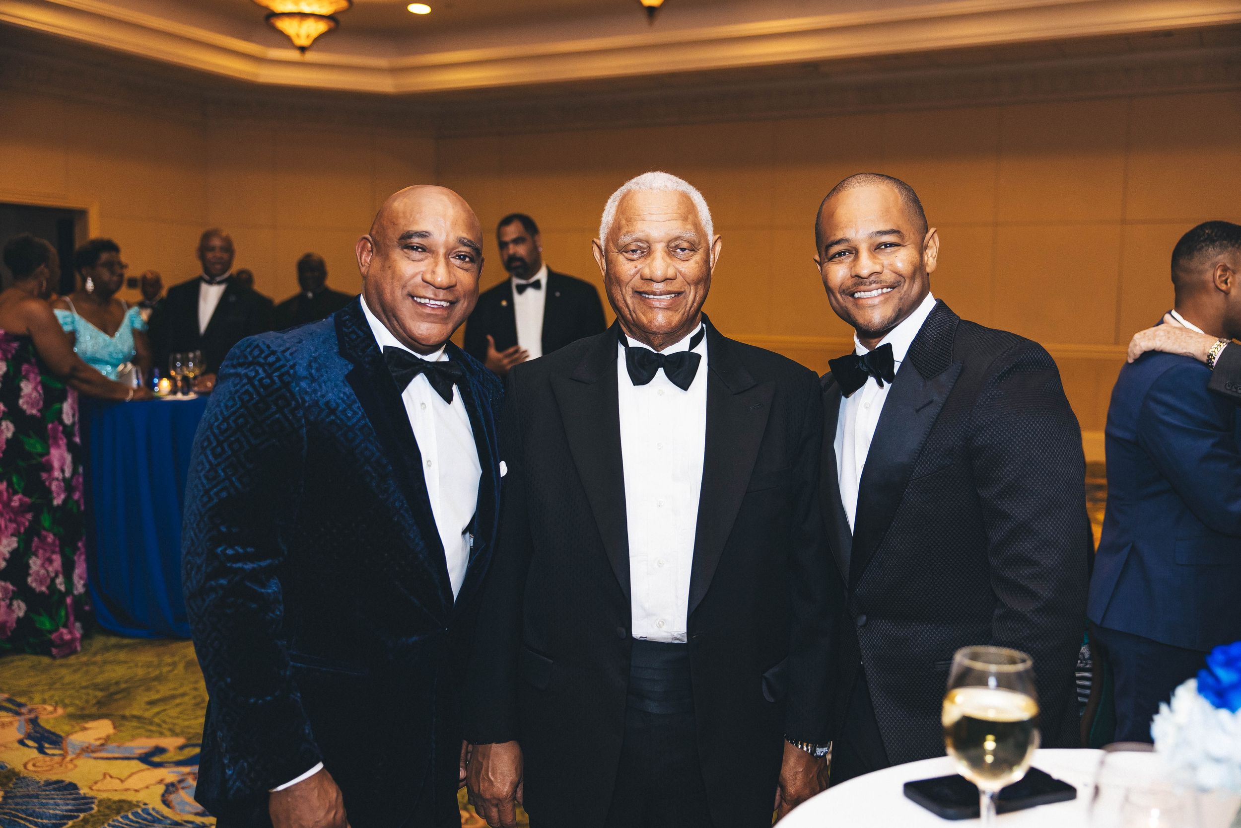 Three men in tuxedos smiling together at a formal gala event in an elegant ballroom setting.