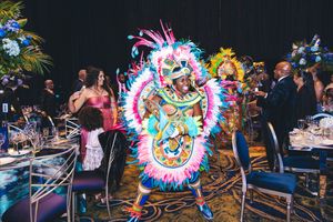 Colorful carnival performer in vibrant feathered costume dancing at an elegant gala event with guests dining in background.