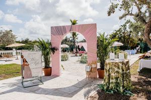 Pink archway entrance to an outdoor wedding venue with tropical palms, patio seating, and elegant table decor on a sunny day.