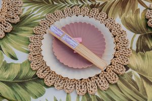 Woven rattan placemat with pink scalloped plates and bamboo chopsticks on tropical leaf tablecloth.