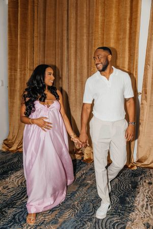 A pregnant woman in a pink gown holds hands with a smiling man in white, standing together in an elegant room.