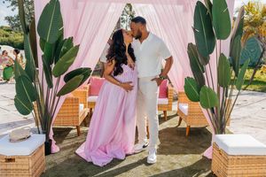 Couple sharing a kiss at an outdoor baby shower under a pink draped canopy with tropical plants.