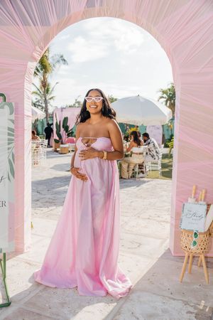 Pregnant woman in pink strapless maternity gown smiling under floral arch at outdoor baby shower event.