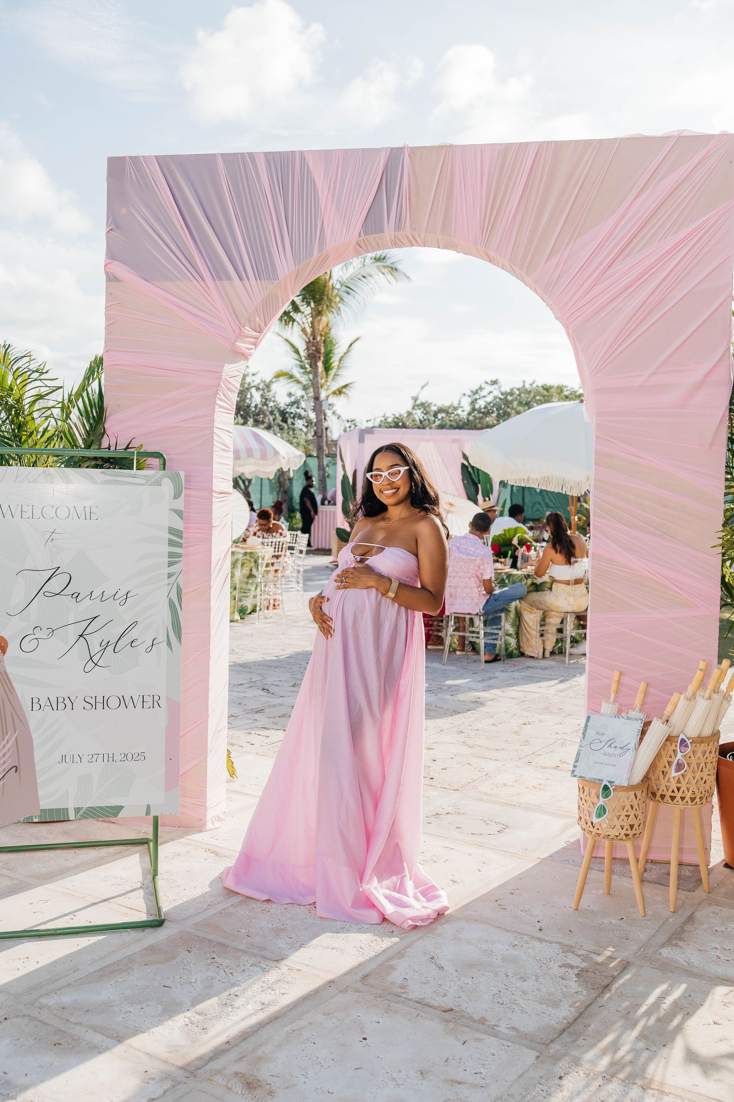 Pregnant woman in pink strapless gown poses under floral arch at elegant outdoor baby shower event.