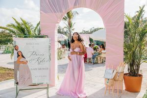 Pregnant woman in pink gown poses under floral arch at elegant outdoor baby shower with tropical decor.