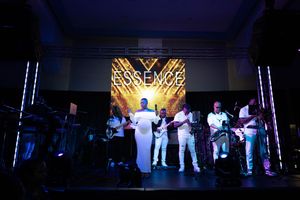 Live band performing on stage at Essence event with glowing branded backdrop and white outfits.