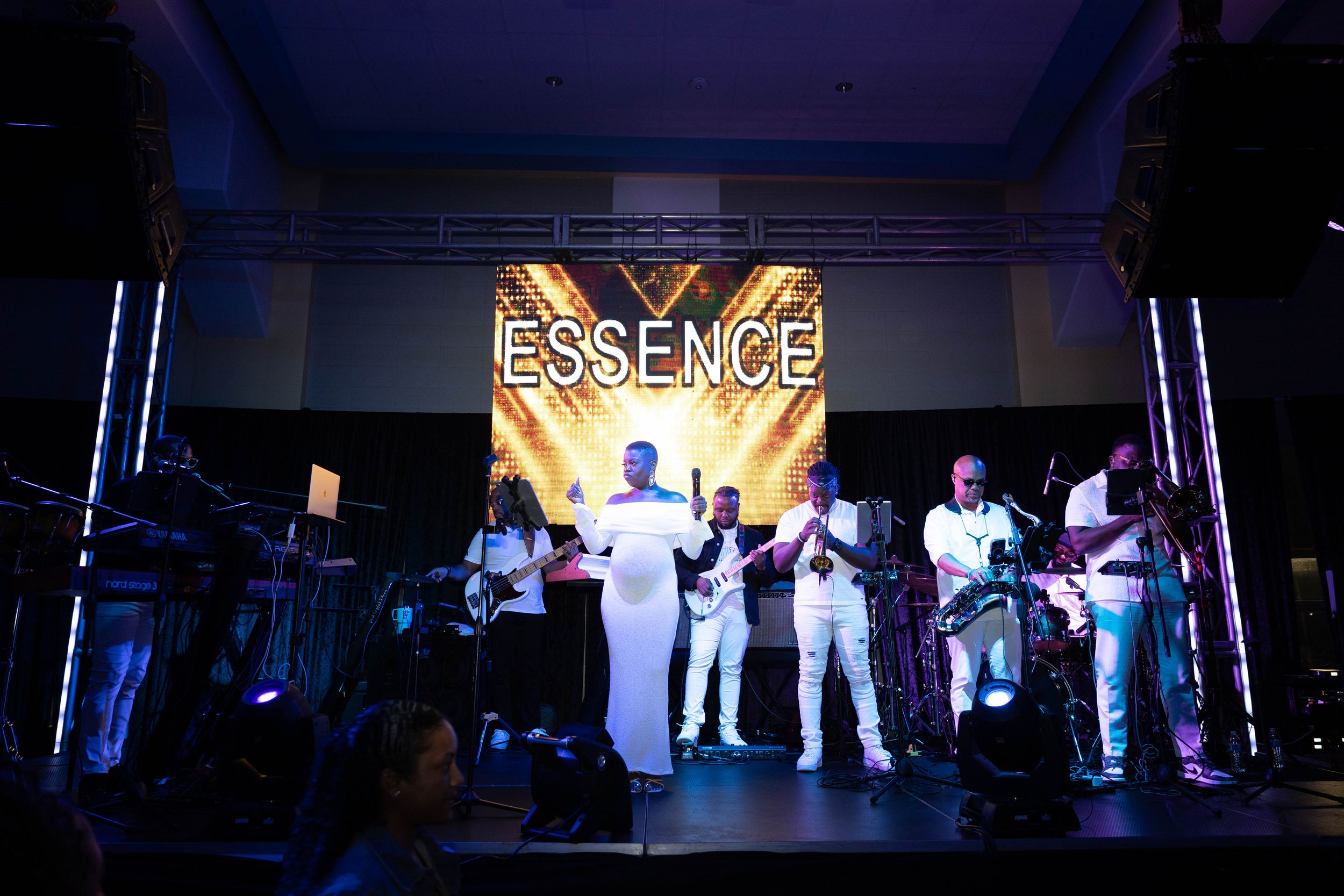 Essence band performing live on stage in white outfits with bright lights and bold signage backdrop.