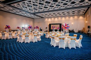 Elegant ballroom with round tables, white chair covers, gold accents, and vibrant floral centerpieces on royal blue carpet.