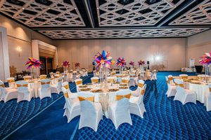 Elegant banquet hall with round tables, white chair covers, gold accents, and colorful feather centerpieces on blue carpet.