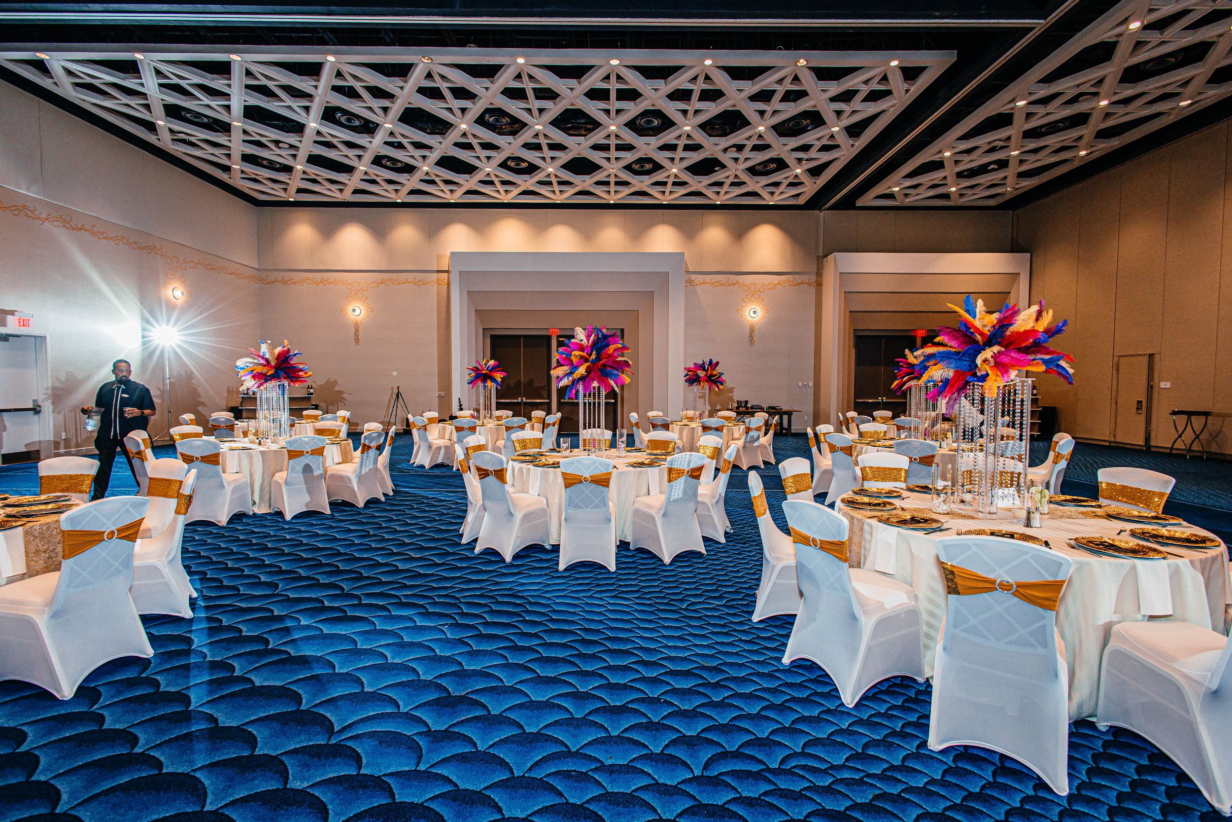 Elegant banquet hall with round tables, white chair covers, colorful floral centerpieces, and a blue quilted carpet.