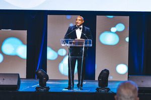 Man in tuxedo speaking at podium on stage with blue bokeh backdrop at formal gala event.