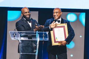 Two men in formal attire on stage, one receiving a framed award certificate at an elegant gala ceremony.