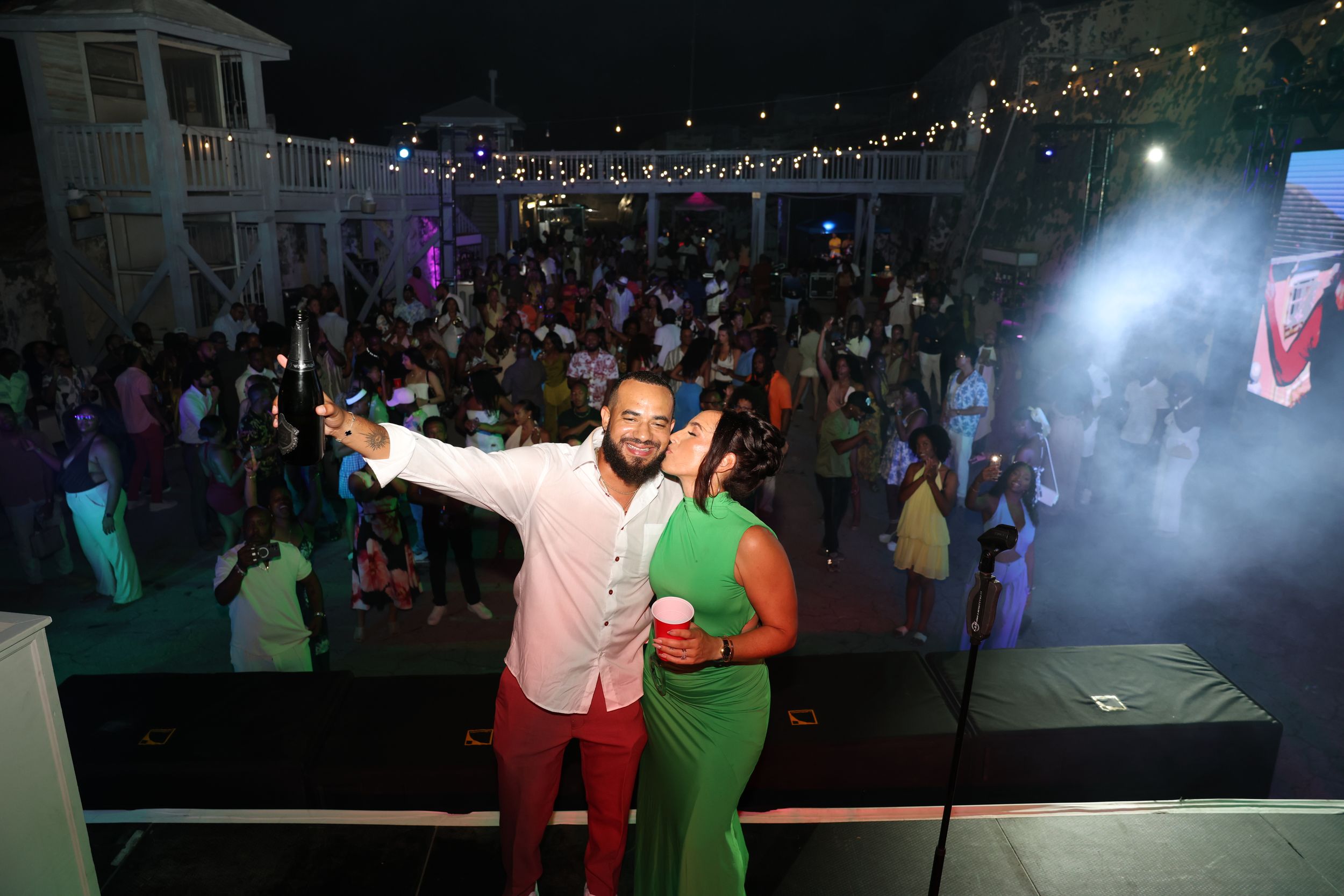 Couple poses on stage at a lively outdoor night party with a large crowd dancing behind them.