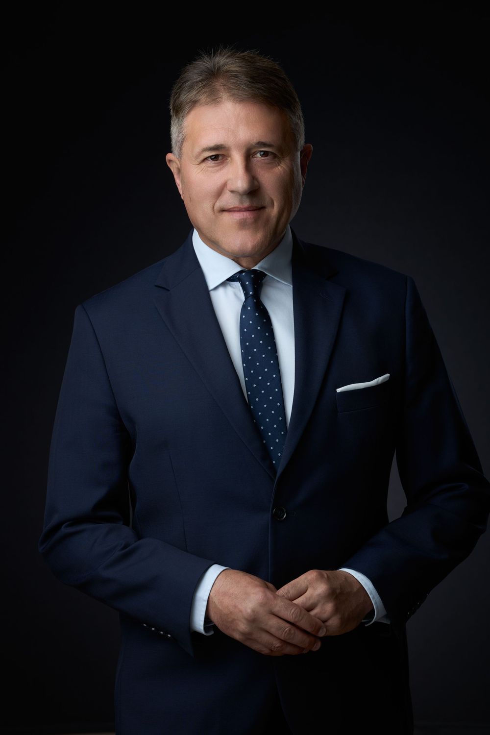 Professional businessman in navy suit and dotted tie poses confidently against dark background in formal portrait.