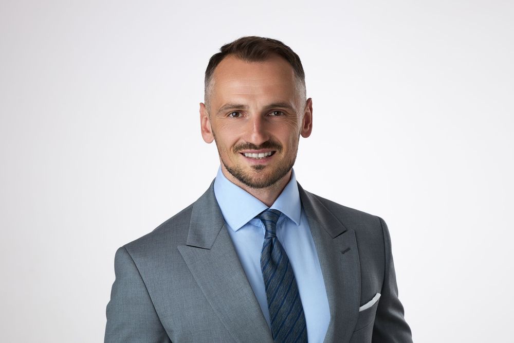 Professional businessman in gray suit and striped tie smiling confidently against white background.