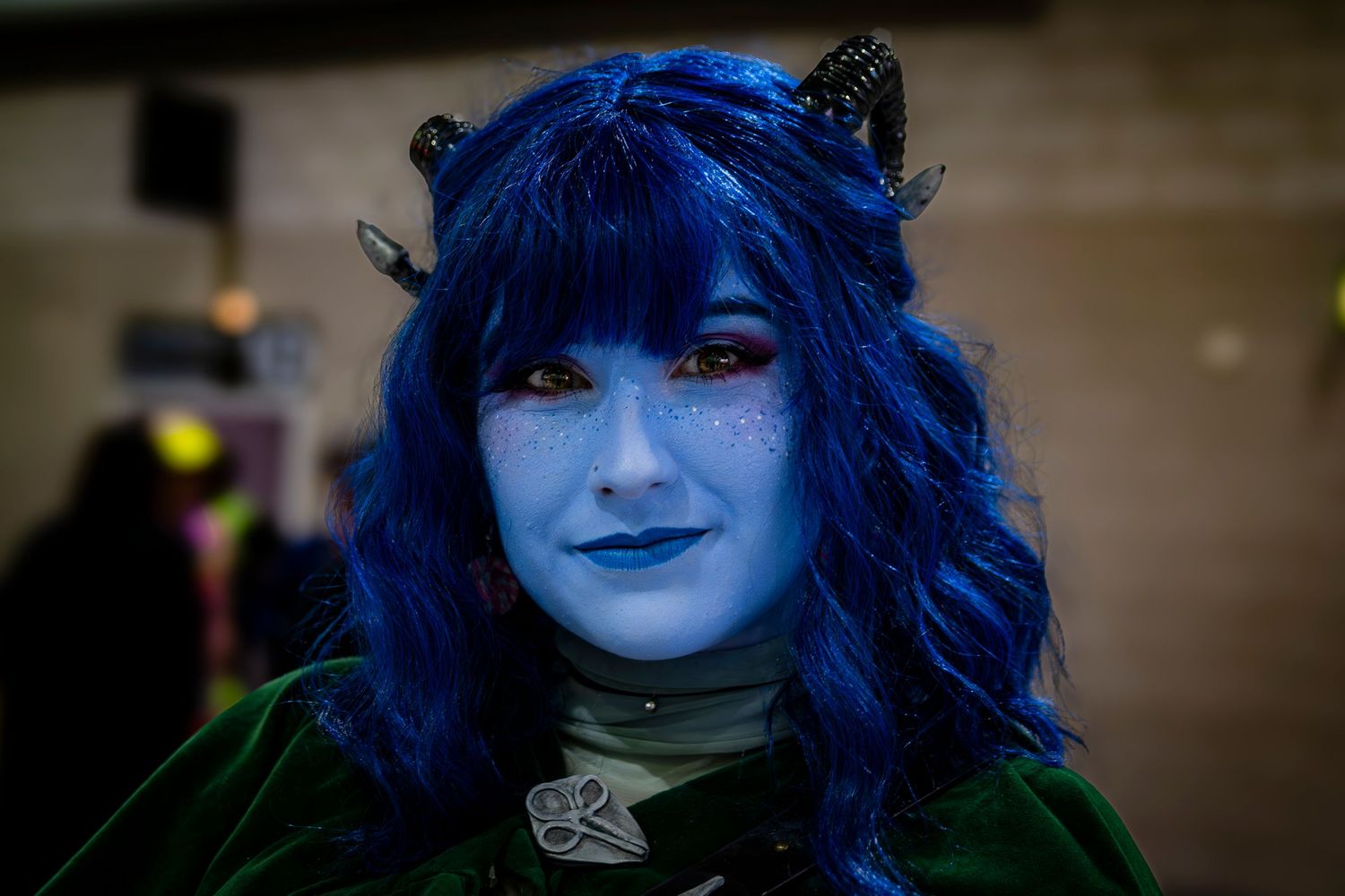 Person in blue fantasy cosplay with horns, blue face paint, and vibrant blue hair at a convention.