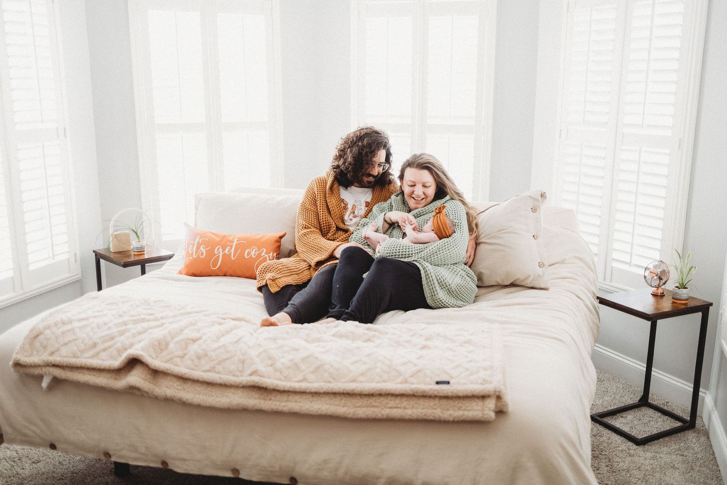 Two women sitting on a cozy bed, wrapped in blankets, smiling and looking at a book together in a bright bedroom.