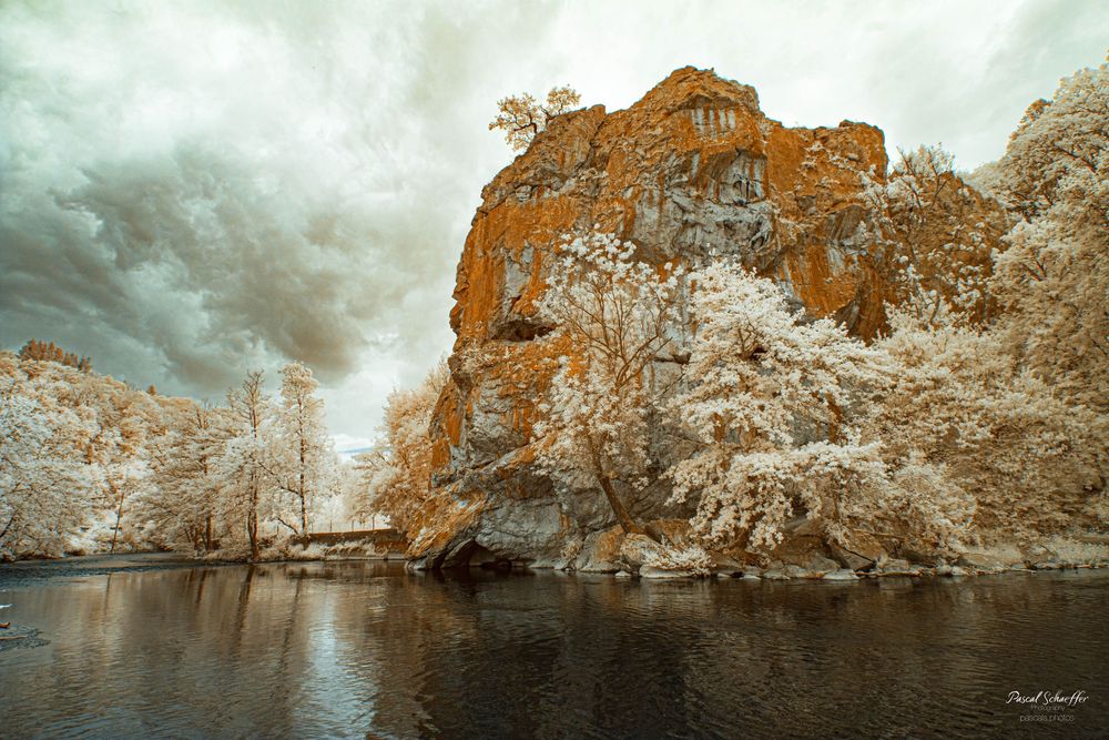 Infrared image of a dramatic orange rocky cliff surrounded by white trees beside a calm river.