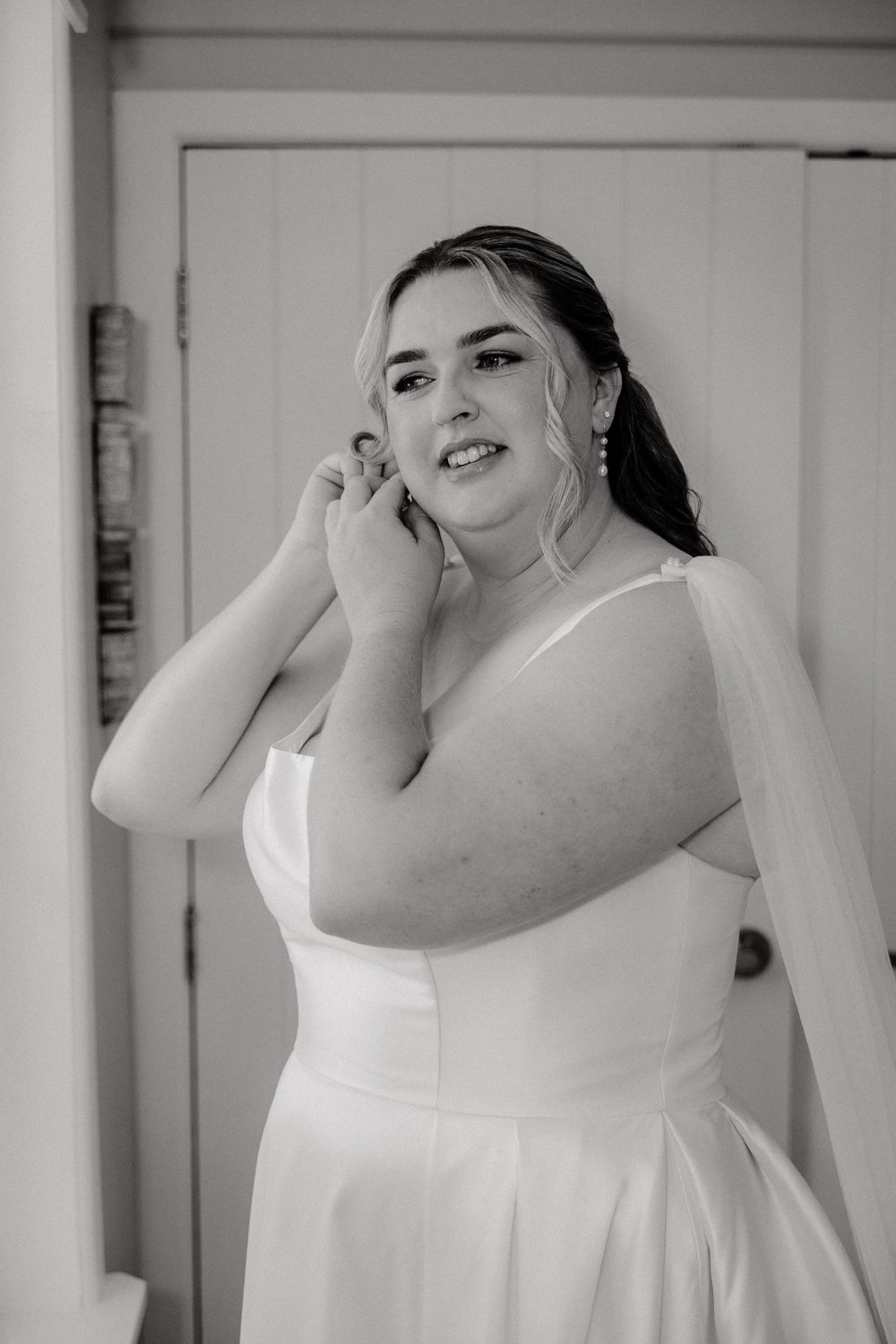 Bride in white satin gown adjusting earrings in black and white bridal portrait.
